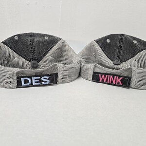 May include: Two black and gray mesh trucker hats with adjustable straps. The hat on the left has the word "DES" embroidered on the front in white. The hat on the right has the word "WINK" embroidered on the front in pink.