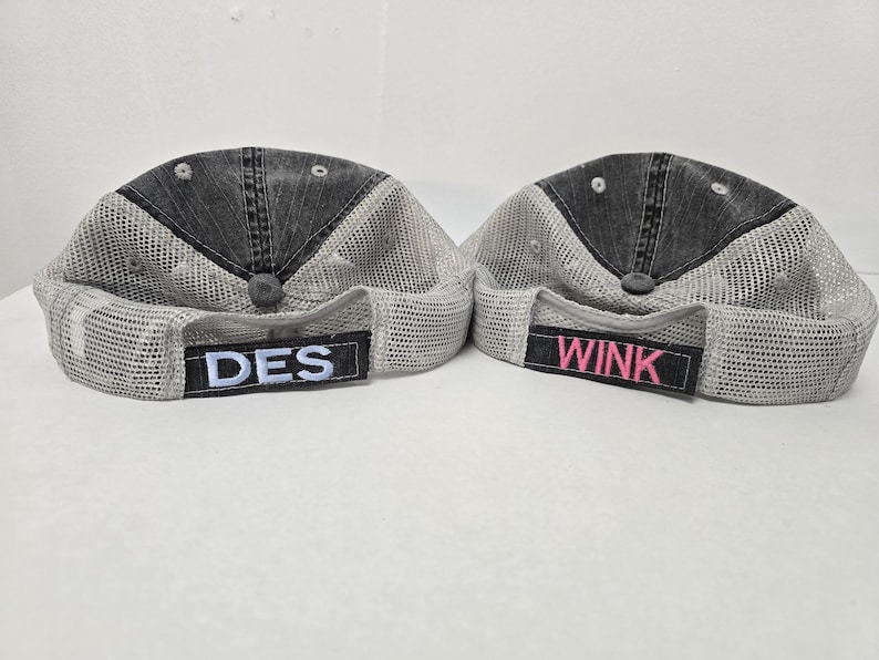 May include: Two black and gray mesh trucker hats with adjustable straps. The hat on the left has the word "DES" embroidered on the front in white. The hat on the right has the word "WINK" embroidered on the front in pink.