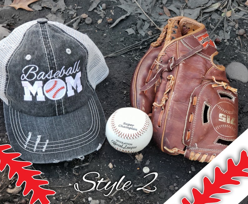 May include: A grey and white baseball cap with the text "Baseball Mom" and a baseball graphic. A brown leather baseball mitt and a white baseball are on a dark brown surface. The text "Style 2" is written at the bottom of the image.