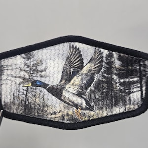 May include: Embroidered patch featuring a flying duck with outstretched wings against a backdrop of a forest scene. The patch has a black border and a detailed design, showcasing the duck's feathers and the trees in shades of gray and black.