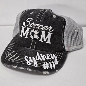 May include: Black and white embroidered trucker hat with the text "Soccer Mom" and a soccer ball graphic. The hat also has the text "Sydney #11" embroidered on the brim.