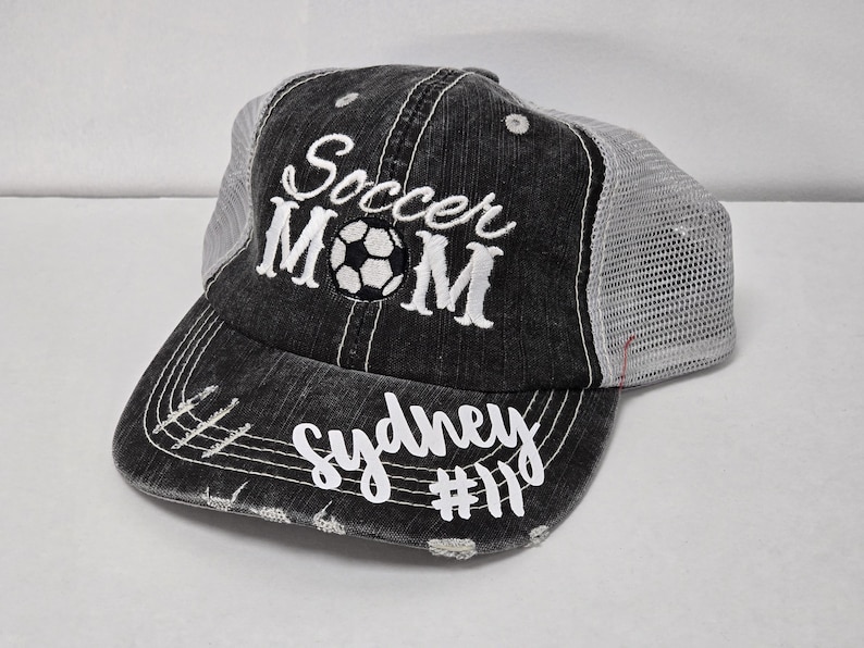 May include: Black and white embroidered trucker hat with the text "Soccer Mom" and a soccer ball graphic. The hat also has the text "Sydney #11" embroidered on the brim.