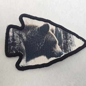 May include: Arrowhead-shaped patch featuring a detailed image of a bear's head and a forest scene. The bear is brown and black, set against a backdrop of trees. The patch has a black border and is likely for clothing or accessories.