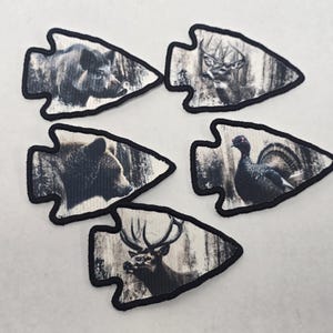 May include: Set of five arrowhead-shaped patches featuring wildlife. The patches depict a bear, deer, elk, and turkey in a forest setting. Each patch has a black border and a neutral color palette.