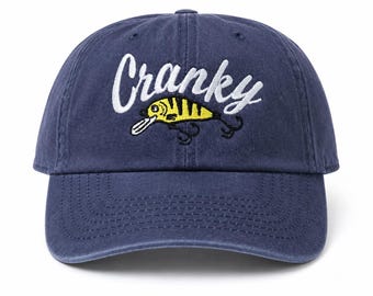 May include: A navy blue baseball cap featuring white embroidered text that reads Cranky above a yellow and black fishing lure graphic with black hooks. The cap has a curved brim and a structured front panel with matching navy blue stitching.