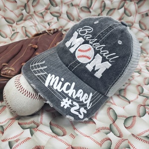 May include: A grey baseball cap with a white embroidered baseball and the words "Baseball Mom" and "Michael #25". The cap is on a white background with a pattern of baseballs.