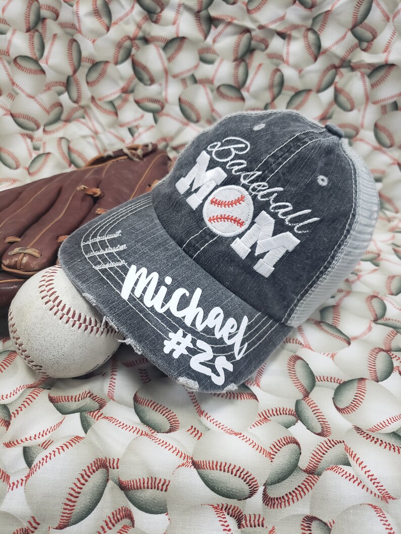 May include: A grey baseball cap with a white embroidered baseball and the words "Baseball Mom" and "Michael #25". The cap is on a white background with a pattern of baseballs.