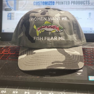May include: A camouflage baseball cap with the text "Women Want Me Fish Fear Me" embroidered on the front. A colorful fish is embroidered below the text.