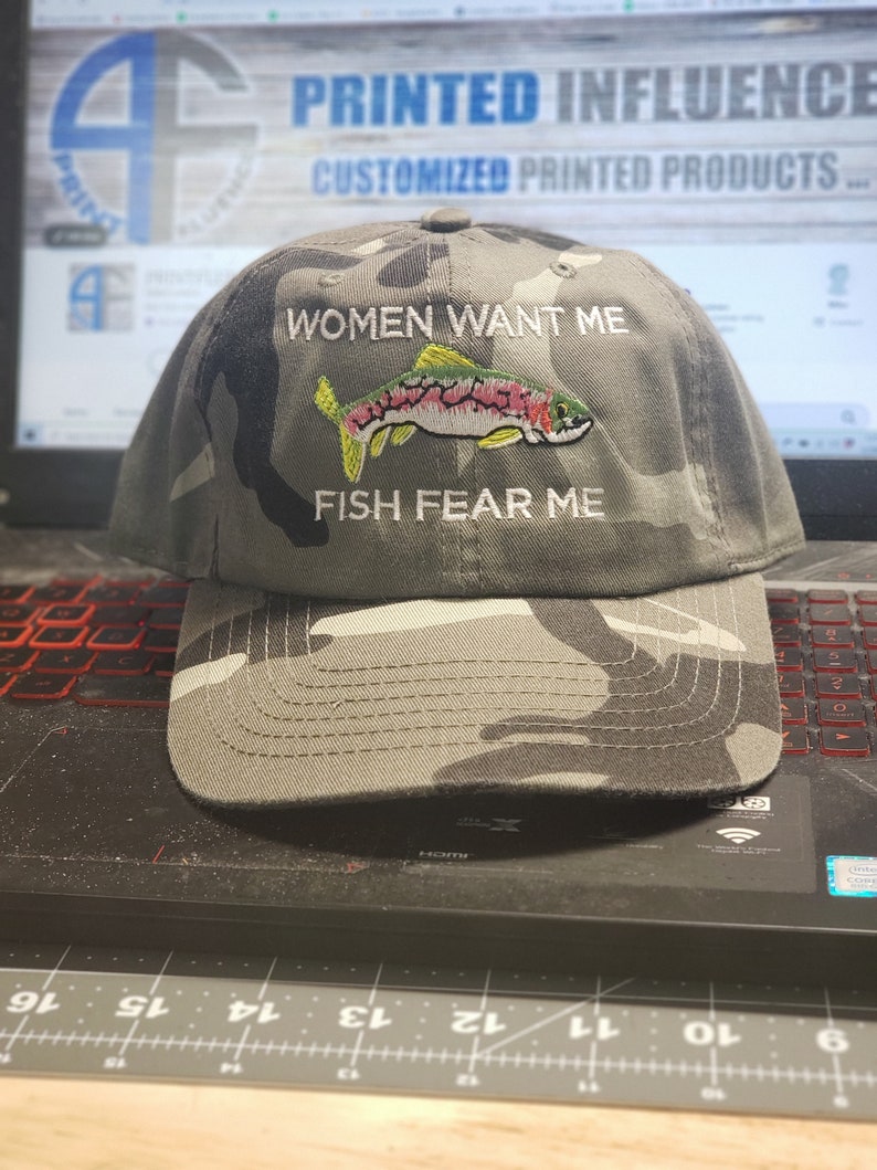 May include: A camouflage baseball cap with the text "Women Want Me Fish Fear Me" embroidered on the front. A colorful fish is embroidered below the text.