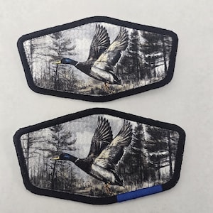 May include: Two embroidered patches featuring a flying duck over a forest scene. The patches have a black border and a blue stripe at the bottom. The duck is depicted with detailed feathers and a yellow beak, set against a grayscale background.