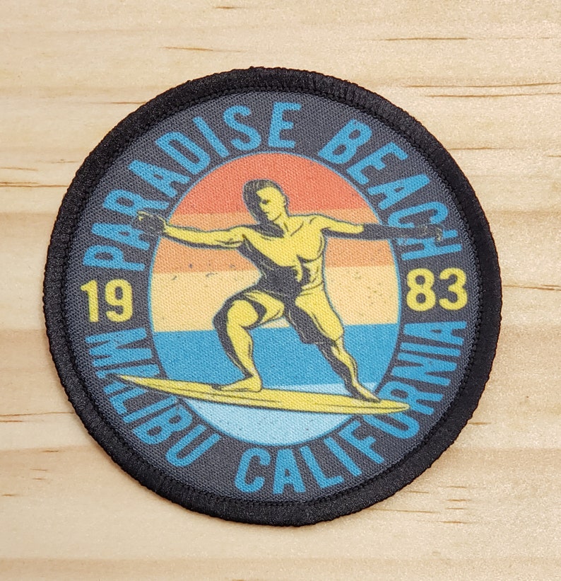 May include: A black and white circular patch with a surfer silhouette in a yellow swimsuit riding a yellow surfboard. The patch has the text "Paradise Beach 1983 Malibu California" in a white, yellow, and orange color scheme.