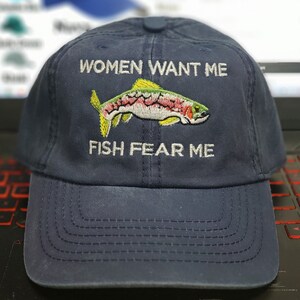 May include: A navy blue baseball cap with white embroidery that reads "Women Want Me Fish Fear Me". The cap features a colorful embroidered trout.