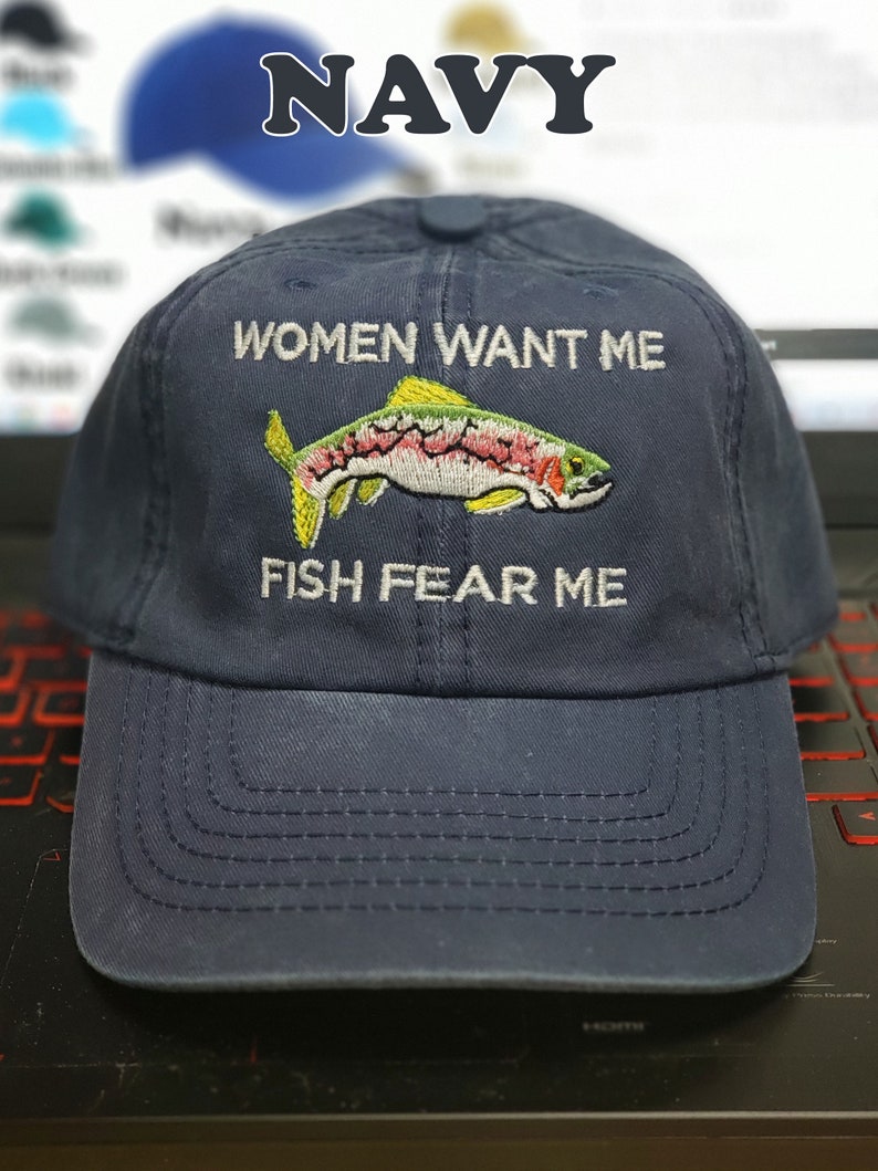 May include: A navy blue baseball cap with white embroidery that reads "Women Want Me Fish Fear Me". The cap features a colorful embroidered trout.
