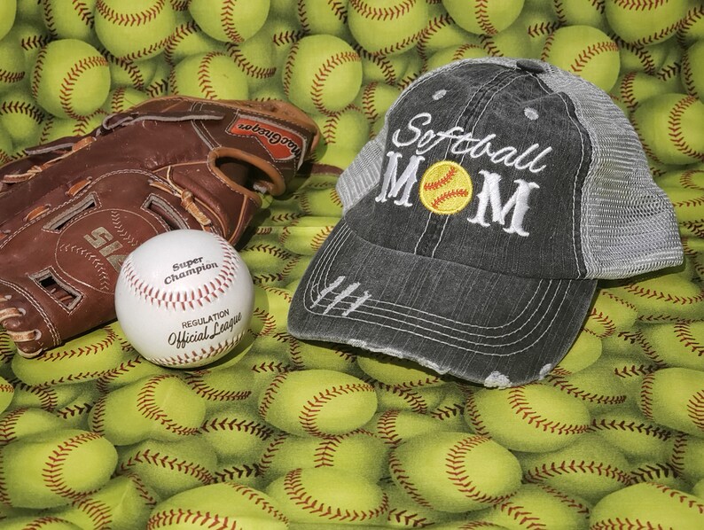 May include: A grey and black baseball cap with the text "Softball Mom" embroidered on the front. A yellow softball with white stitching is embroidered inside the word "Mom". The cap is surrounded by yellow baseballs.