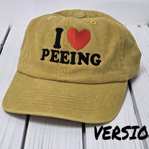 May include: A khaki baseball cap with black embroidery that reads "I ♥ PEEING" with a red heart.