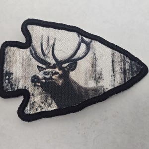 May include: Arrowhead-shaped patch featuring a detailed image of a majestic elk with large antlers set against a grayscale forest backdrop. The patch has a black border, adding a defined edge to the design. Ideal for outdoor enthusiasts.