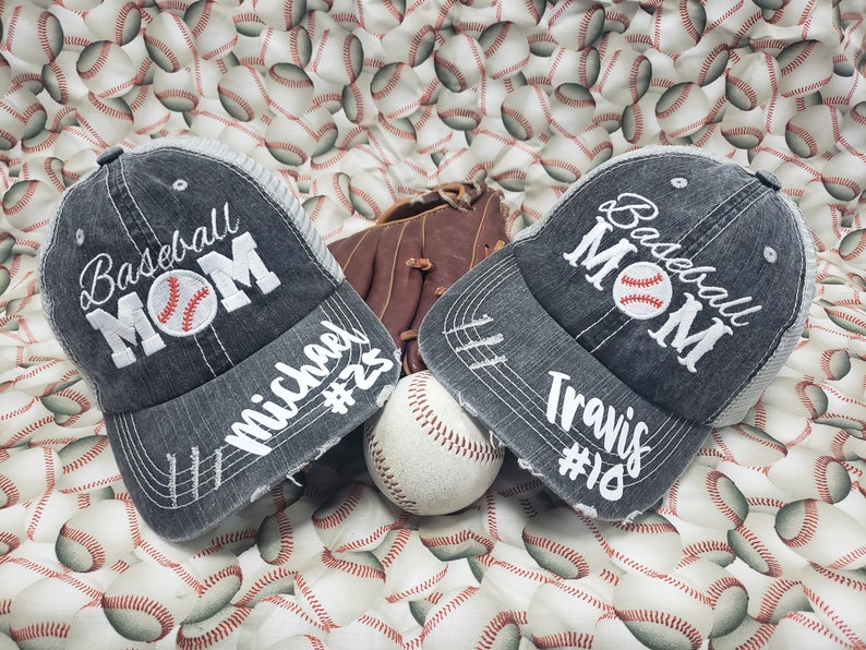 May include: Two grey baseball caps with the text "Baseball Mom" embroidered on the front. The caps have white mesh panels and are decorated with white stitching. One cap has the name "Michael #25" written on the side, and the other has the name "Travis #18".