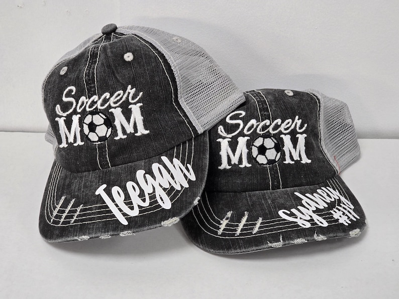 May include: Two black and gray trucker hats with white embroidered text that reads "Soccer Mom" and a soccer ball. The hats have a white mesh back and the text "Tegan" and "Sydney #11" are embroidered on the front of each hat.