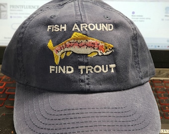 May include: Navy blue baseball cap with the embroidered text "FISH AROUND FIND TROUT" and a colorful fish graphic. The hat has a curved brim and a button on top. A perfect accessory for fishing enthusiasts.