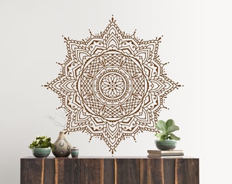 May include: A large, brown mandala wall decal with intricate patterns is displayed on a white wall. The decal is above a dark wood cabinet with metal legs. Decorative vases and plants are on the cabinet.