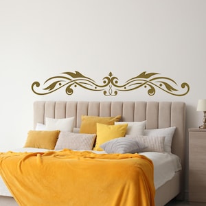 May include: A gold-coloured wall decal with a symmetrical, flowing design. The decal features swirling lines, dots, and a central flourish. It is positioned above a bed with a beige headboard and yellow bedding, creating a decorative bedroom feature.