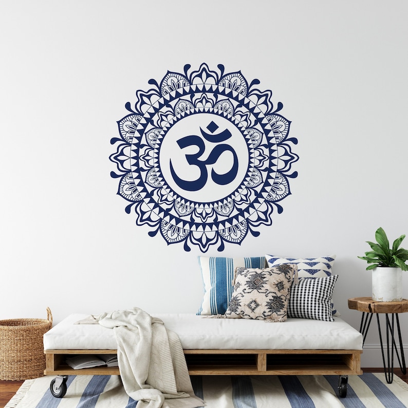 May include: A navy blue Om symbol mandala wall decal with intricate details. The decal is displayed on a white wall above a white couch with pillows and a throw blanket.