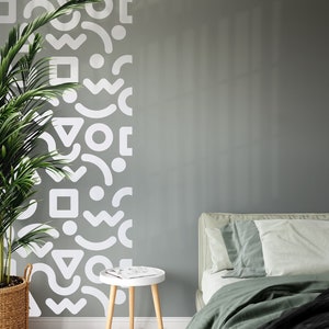 Op de afbeelding: Een slaapkamer met een groene bedsprei en witte geometrische vormen op de muur. Een klein houten bijzettafeltje met een tijdschrift erop staat naast het bed.
