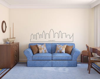 Könnte beinhalten: Wohnzimmer mit blauem Sofa, Kissen, Holzschrank und Wandkunst mit der Skyline von Cincinnati. Der Raum hat einen hellblauen Teppich und einen Schreibtisch mit einem Stuhl.