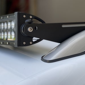 May include: A black LED light bar with multiple bright lights, mounted on a vehicle. The light bar is attached to a black bracket, secured with bolts. The vehicle's surface is white.