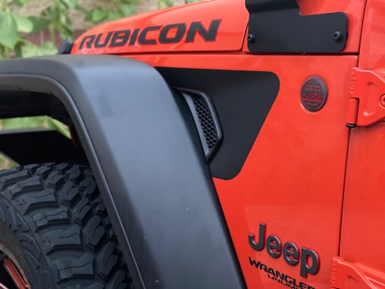 May include: A close-up of a bright orange Jeep Wrangler Rubicon with black fender flares and a black Jeep logo. The Jeep has a black grill and black wheels with all-terrain tires.