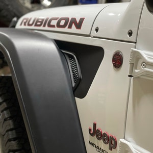 May include: A white Jeep Wrangler Rubicon with black fender flares and red accents. The Jeep has a black grille and a black bumper. The Jeep is parked in a garage.