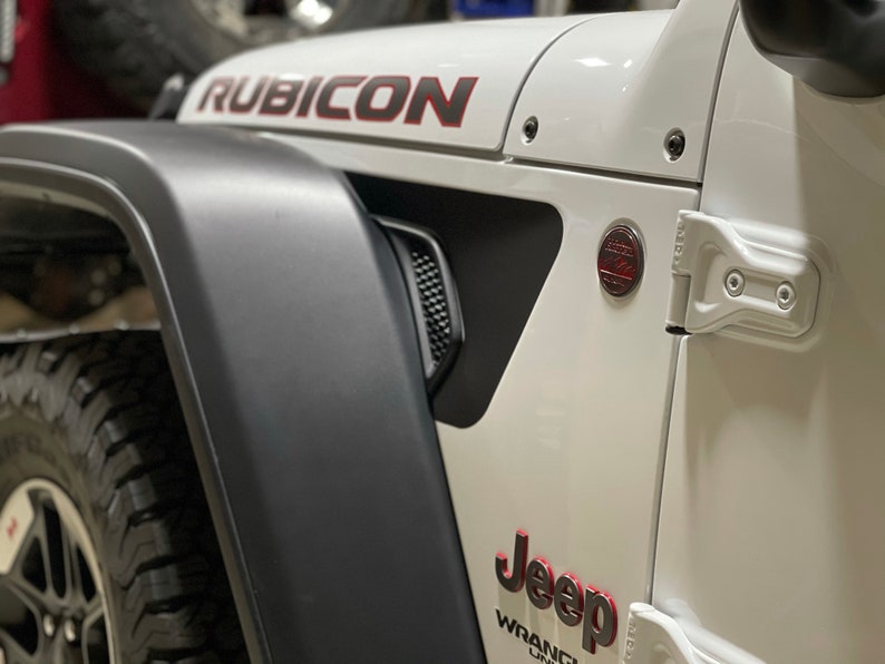 May include: A white Jeep Wrangler Rubicon with black fender flares and red accents. The Jeep has a black grille and a black bumper. The Jeep is parked in a garage.