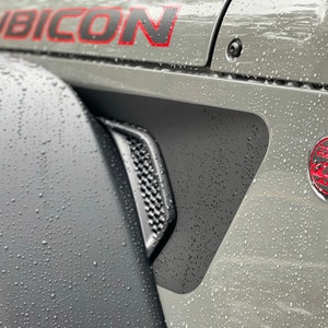 May include: A gray Jeep Rubicon with a black and red "Trail Rated" badge on the side. The vehicle is covered in water droplets.