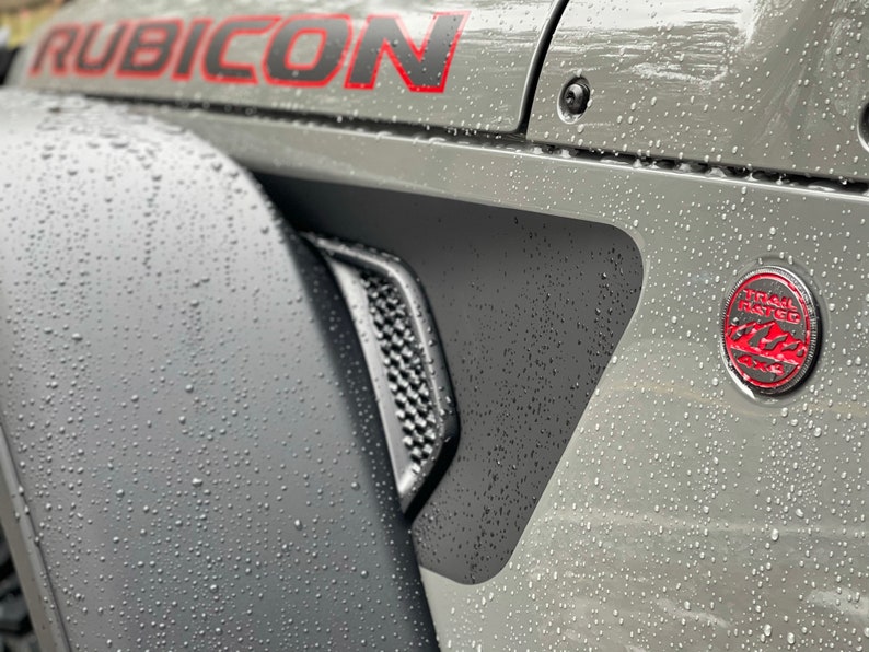 May include: A gray Jeep Rubicon with a black and red "Trail Rated" badge on the side. The vehicle is covered in water droplets.