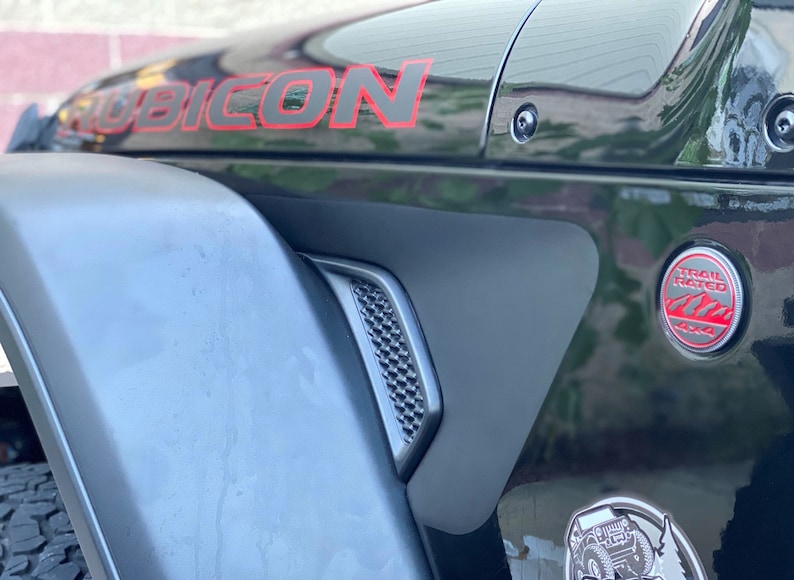 May include: A black Jeep Wrangler Rubicon with red accents. The vehicle has a Trail Rated badge and a Jeep logo on the fender.