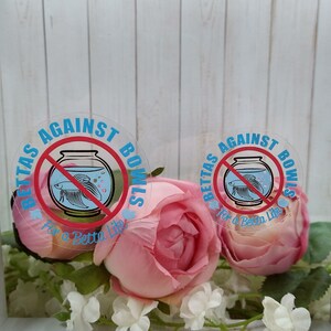 May include: Two round stickers with a blue and red betta fish inside a bowl with a red circle and line through it. The text on the stickers reads "BETTAS AGAINST BOWLS For a Betta Life". The stickers are on a white background with pink flowers.