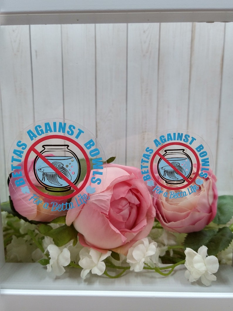 May include: Two round stickers with a blue and red betta fish inside a bowl with a red circle and line through it. The text on the stickers reads "BETTAS AGAINST BOWLS For a Betta Life". The stickers are on a white background with pink flowers.