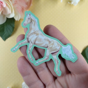 May include: A white horse sticker with a light green outline. The horse is in a running pose with its head turned to the side. The sticker is held in a hand.
