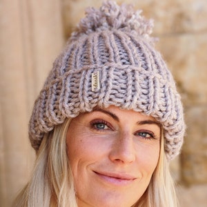 May include: A light brown knitted beanie with a fluffy pom pom on top. The beanie has a ribbed texture and a small label that says "dashi" on the side.