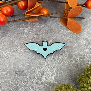 May include: A light blue enamel pin shaped like a bat with a small black heart in the center. The pin is on a gray background.