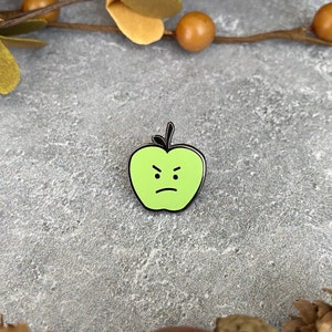 May include: A green apple enamel pin with a black outline and a frowning face.
