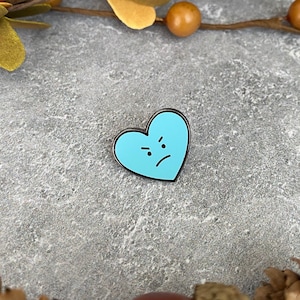 May include: A blue enamel pin shaped like a heart with a frowning face. The pin is on a gray background.
