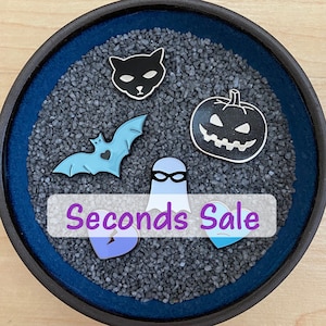 May include: A collection of six enamel pins in a blue bowl filled with black sand. The pins feature a black cat, a ghost with a mask, a bat, a pumpkin, a broken heart, and a blue heart. The text "Seconds Sale" is written in purple on a white banner.