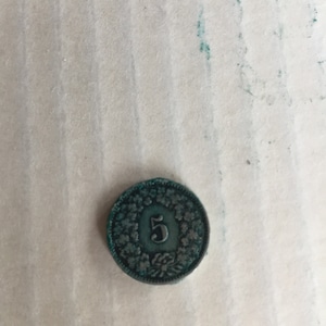 May include: A close-up of a green metal coin with a wreath design and the number "5" in the center.