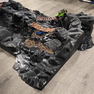 以下が含まれることがあります： 橋、トンネル、さまざまな岩石層を備えた、詳細な黒い地形モデル。 2つの小さな玩具車（オレンジと緑）がモデル上に配置されており、ミニチュアオフロードアドベンチャーのセットアップを示唆しています。