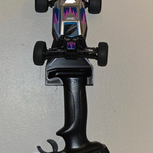Peut inclure: Un support mural pour ranger une voiture télécommandée et sa manette. La voiture est argentée avec des accents rose et bleu et un aileron arrière transparent. La manette est noire avec une gâchette et un volant.