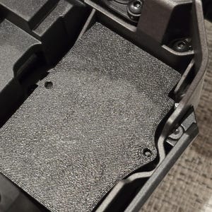 May include: A close-up of a black plastic component with a textured surface. The component has a rectangular shape and is attached to a larger black plastic object.