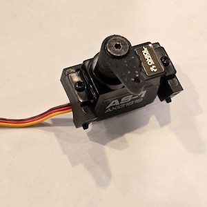 以下が含まれることがあります： 銀色の AS-1 AX131619 ラベルが付いた黒いサーボモーター。サーボは黒いプラスチックアームと小さな銀色のネジが付いています。サーボは赤、黄、黒のワイヤーが付いた3芯ケーブルに接続されています。