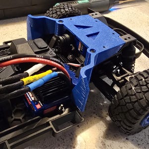 May include: A partially assembled blue and black remote control truck chassis. The truck features a blue 3D-printed component, black tires, and various electronic components, including wires and a motor.