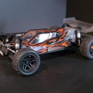 May include: A grey and orange remote control car with black wheels and a clear spoiler. The car has a detailed design with orange accents and a clear windscreen. The tyres have a rugged tread pattern.